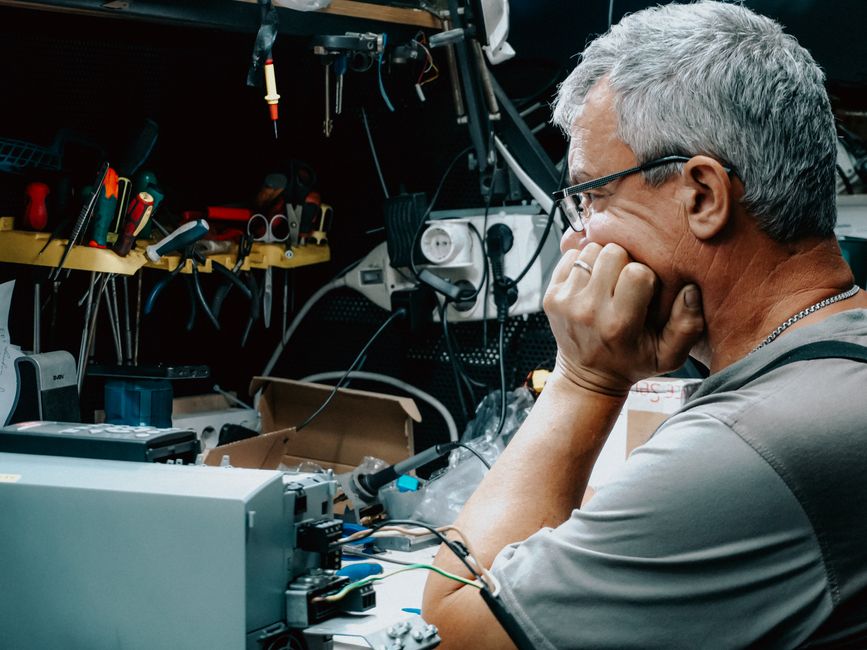 Odrasli muškarac pregledava elektroničku opremu na pretrpanom radnom stolu, zadubljen u razmišljanje. Popravak elektronike u unutarnjem prostoru.